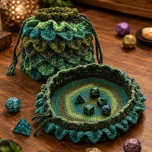 Puede incluir: Una bolsa de ganchillo con cordón en tonos verde y azul cerceta con textura de escamas y una bandeja circular de ganchillo a juego con borde ondulado. Varios dados de juego de múltiples caras de colores están esparcidos por el lugar.