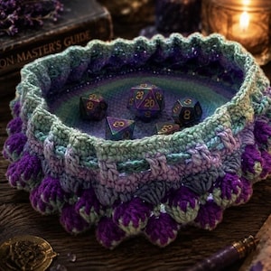 May include: A round, crocheted bowl featuring a gradient of purple, teal, and light green yarn. The exterior has a textured, bobble-stitch pattern, while the interior holds five multi-faceted polyhedral dice in shades of dark purple and gold.