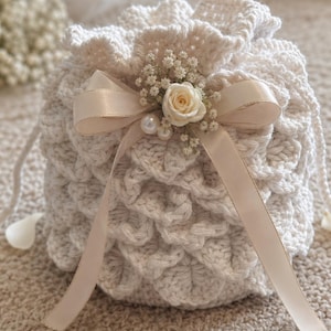 Può includere: Un sacchetto a coulisse all'uncinetto color crema con un motivo strutturato. La borsa è adornata con un nastro color crema, una piccola rosa bianca, delicati fiori bianchi e due perle. Ideale per un matrimonio o un'occasione speciale.
