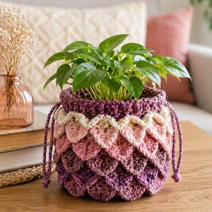 May include: A crocheted plant pot with a gradient of pink and purple scales, holding a green leafy plant. The pot has a drawstring closure and sits on a wooden table. A small glass vase with dried flowers is in the background.