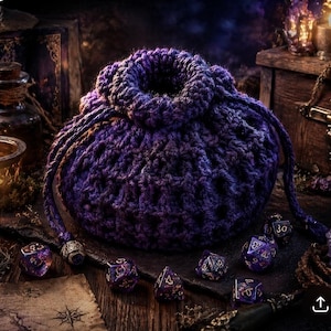 Peut inclure: Un sac à dés en crochet violet foncé fait à la main, avec une fermeture à cordon. Plusieurs dés polyédriques violets et dorés sont disposés autour du sac, suggérant un thème de jeu de société. L'arrière-plan comprend un coffre au trésor et une bougie allumée.