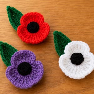 Puede incluir: Tres broches de flores tejidos a ganchillo en colores rojo, morado y blanco, cada uno con un centro negro y una hoja verde. Estos accesorios textiles hechos a mano presentan un acabado de hilo texturizado y una estructura de pétalos superpuestos.