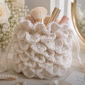 May include: A cream-colored, crocheted drawstring bag with a textured, scale-like pattern. The bag is filled with makeup brushes and tubes. The bag is approximately 6 inches tall and 5 inches wide.