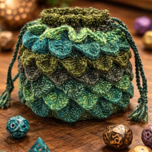 Könnte beinhalten: Ein handgefertigter, grün-blauer gehäkelter Würfelbeutel mit Kordelzug und Quasten. Die Tasche hat eine schuppenartige Textur und ist von verschiedenen polyedrischen Würfeln auf einer Holzoberfläche umgeben. Die Tasche ist etwa 13 cm hoch.