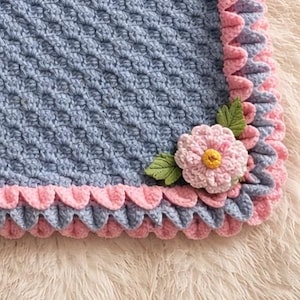 Puede incluir: Manta de bebé tejida a ganchillo en un patrón azul texturizado con un borde rosa festoneado. Una gran flor rosa con centro amarillo y hojas verdes está adjunta. Suave y hecha a mano.