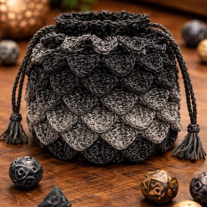 May include: A handmade, crocheted dice bag with a dragon scale design. The bag has a drawstring closure with tassels and a colour gradient from dark grey to light grey. The bag is on a wooden surface with several dice.