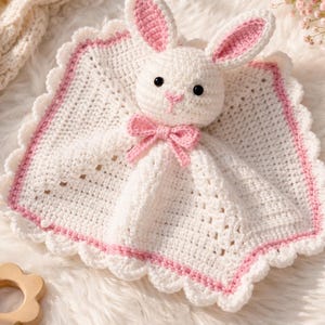 May include: A handmade white crochet bunny lovey with pink accents. The bunny has black eyes, pink inner ears, and a pink bow. The blanket has a scalloped edge with a pink border. The lovey is on a white, fluffy surface.