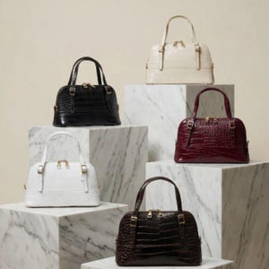 Puede incluir: Seis bolsos con forma de cúpula en varios colores: negro, blanco, crema, burdeos y marrón. Cada bolso presenta un patrón texturizado similar al de un cocodrilo, asas superiores y herrajes dorados. Los bolsos se exhiben sobre pedestales de bloques de mármol.