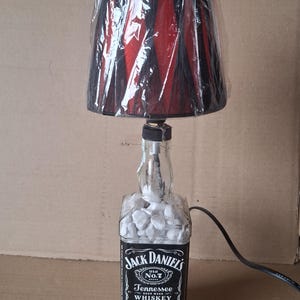 Può includere: Una lampada da tavolo unica realizzata con una bottiglia di whisky Jack Daniel's. La lampada presenta un paralume nero e rosso ed è riempita di piccole pietre bianche. L'etichetta della bottiglia è ben visibile.
