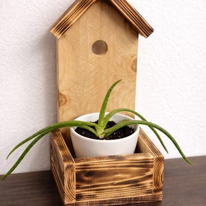 May include: A wooden birdhouse-shaped planter with a small round hole and a rectangular base. A white ceramic pot with a green plant is inside the base. The wood has a natural finish with darker, burnt accents.