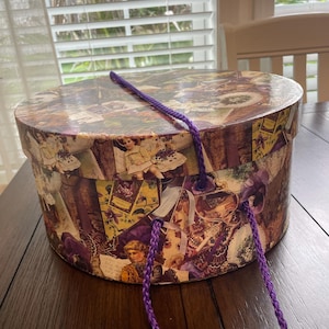 May include: A round cardboard hat box featuring a collage of vintage-style illustrations, including portraits of children and floral motifs in shades of purple, yellow, and brown. The box includes a matching lid and a braided purple rope handle.