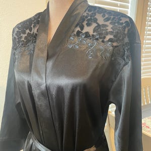 May include: Black satin robe with a shawl collar and a matching tie belt. The robe features sheer floral lace detailing on the shoulders and upper back. The sleeves are three-quarter length, and the robe is displayed on a mannequin.