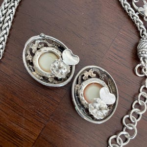 May include: Silver-toned oval earrings with a cream-colored center, a heart, and a flower accent. A silver chain necklace is also visible. The earrings and necklace are on a brown surface.