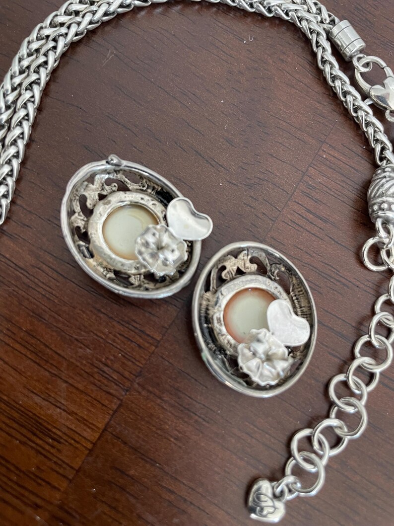 May include: Silver-toned oval earrings with a cream-colored center, a heart, and a flower accent. A silver chain necklace is also visible. The earrings and necklace are on a brown surface.