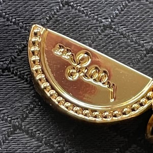 May include: Close-up of a gold-colored pendant with a semi-circular shape. The pendant has a beaded border and an embossed design with the word "Forever" in cursive script. The background is a textured black fabric.