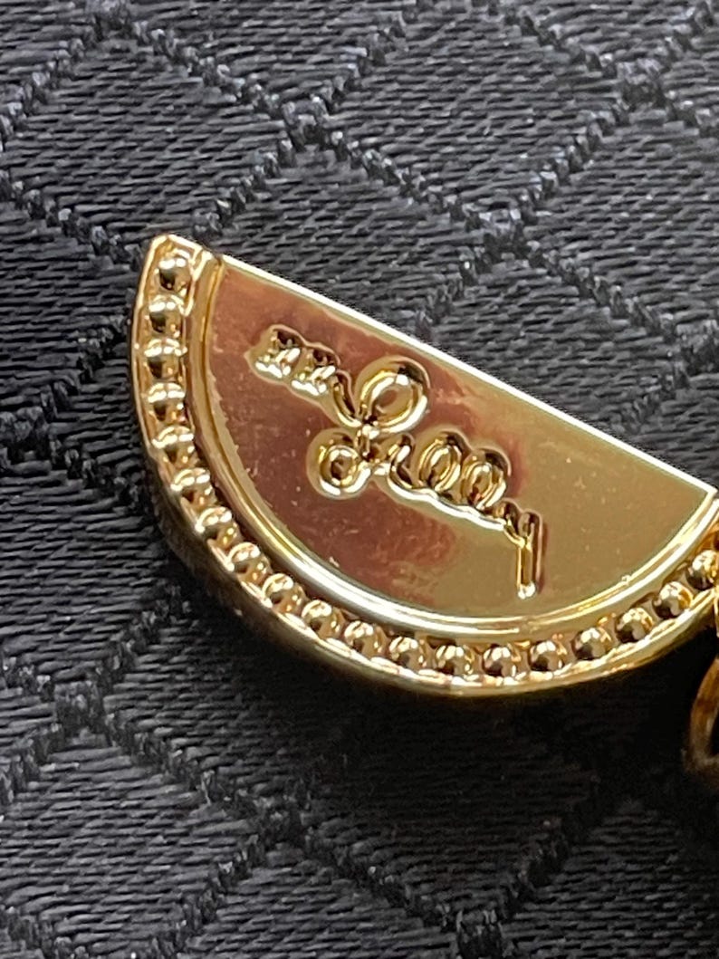May include: Close-up of a gold-colored pendant with a semi-circular shape. The pendant has a beaded border and an embossed design with the word "Forever" in cursive script. The background is a textured black fabric.