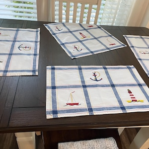 May include: Four nautical-themed placemats on a dark brown wooden table. Each white placemat features a blue plaid pattern and various nautical illustrations, including anchors, sailboats, and lighthouses. The placemats are rectangular.