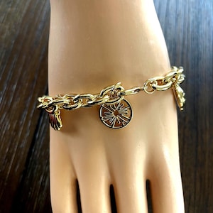 May include: Gold-toned charm bracelet featuring a chain link design. The bracelet has several charms, including a sunburst design, a seashell, and a small decorative element. The bracelet is displayed on a mannequin hand.