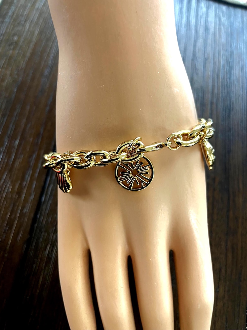 May include: Gold-toned charm bracelet featuring a chain link design. The bracelet has several charms, including a sunburst design, a seashell, and a small decorative element. The bracelet is displayed on a mannequin hand.