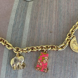 May include: Gold-toned charm bracelet with various charms, including a seashell, elephant, and a pink floral design. Other charms include a lemon slice and a decorative wheel. The bracelet is on a striped background.