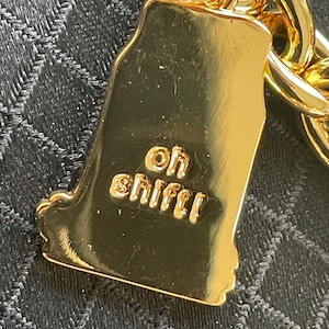 May include: A gold-colored charm in the shape of a state, with the words "oh sh!ft!" embossed on it. The charm is attached to a gold chain. The background is a dark, textured fabric.