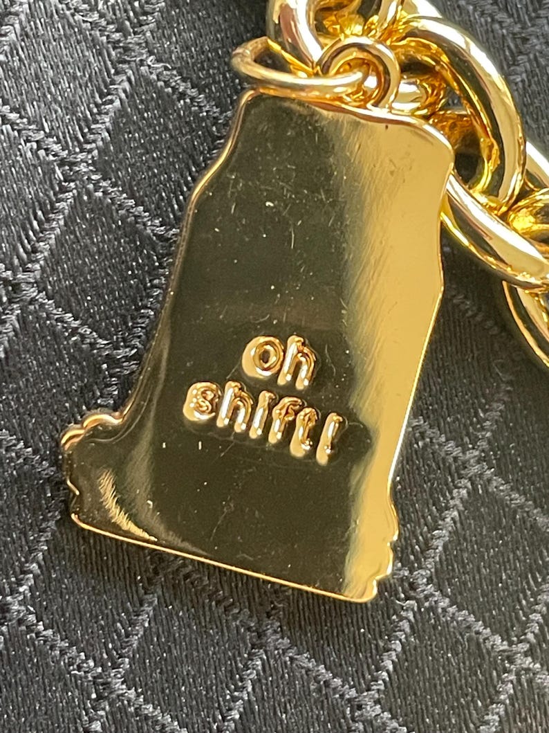 May include: A gold-colored charm in the shape of a state, with the words "oh sh!ft!" embossed on it. The charm is attached to a gold chain. The background is a dark, textured fabric.