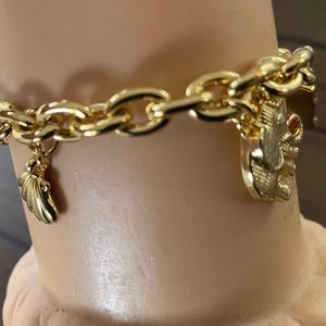 May include: A gold-colored chain bracelet with multiple charms. The bracelet features a chunky chain design with a wing charm and a charm with an elephant design. The bracelet is displayed on a mannequin arm.