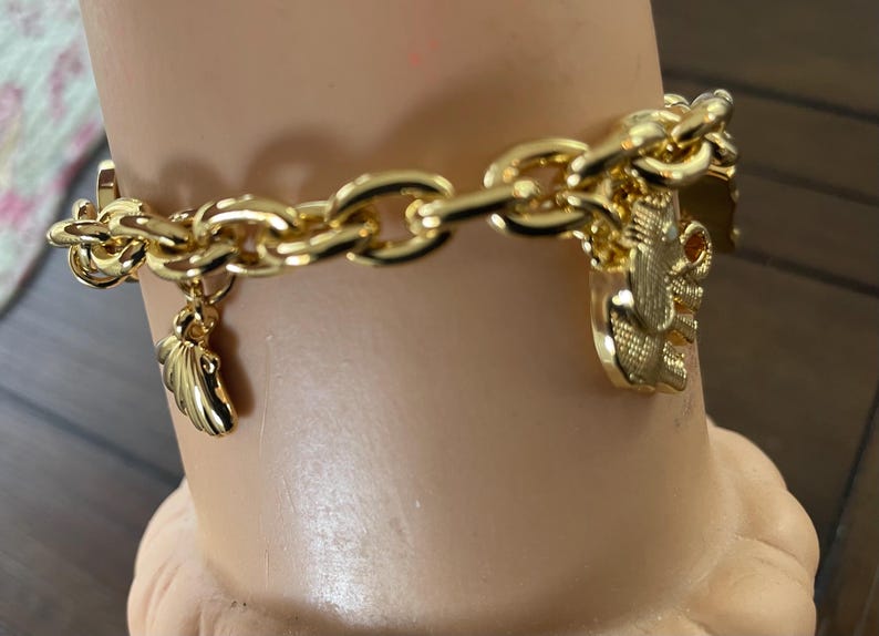 May include: A gold-colored chain bracelet with multiple charms. The bracelet features a chunky chain design with a wing charm and a charm with an elephant design. The bracelet is displayed on a mannequin arm.