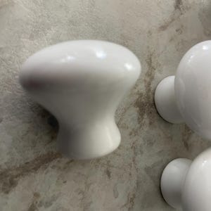 May include: White ceramic cabinet knobs with a rounded top and a flared base. The knobs have a glossy finish and are set against a light-colored, textured background. These knobs are suitable for drawers and cabinets.