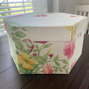 May include: A hexagonal storage box with a floral pattern. The box features a white background with watercolor-style flowers in shades of pink, yellow, and green. The box is sitting on a dark brown surface.