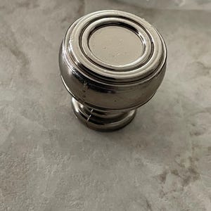 May include: A silver-toned cabinet knob with a rounded top and a circular design. The knob has a short, cylindrical base and is likely made of metal. The finish appears to be brushed or polished, giving it a modern look.