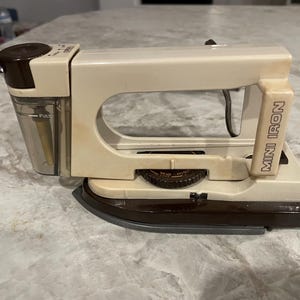 May include: A cream-colored mini iron with a brown base and handle. The iron has a water reservoir and a dial with settings. The words "MINI IRON" are printed on the side.