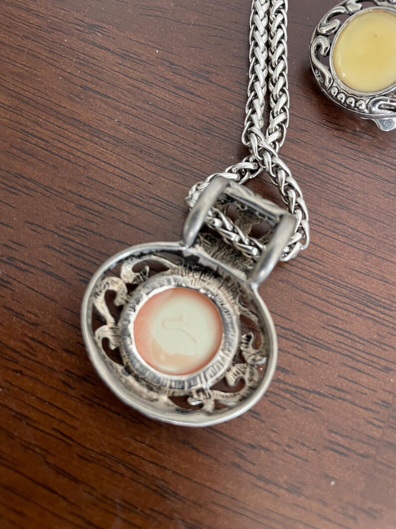 May include: Silver-toned necklace with a pendant featuring a round, peach-colored center. The pendant has an ornate, openwork design around the center. A matching silver-toned earring with a yellow center is also visible.