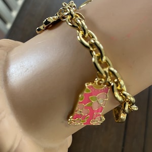 May include: Gold-toned charm bracelet with a chunky chain. The bracelet features several charms, including a pink and green floral-decorated charm. The bracelet is on a light-colored surface.