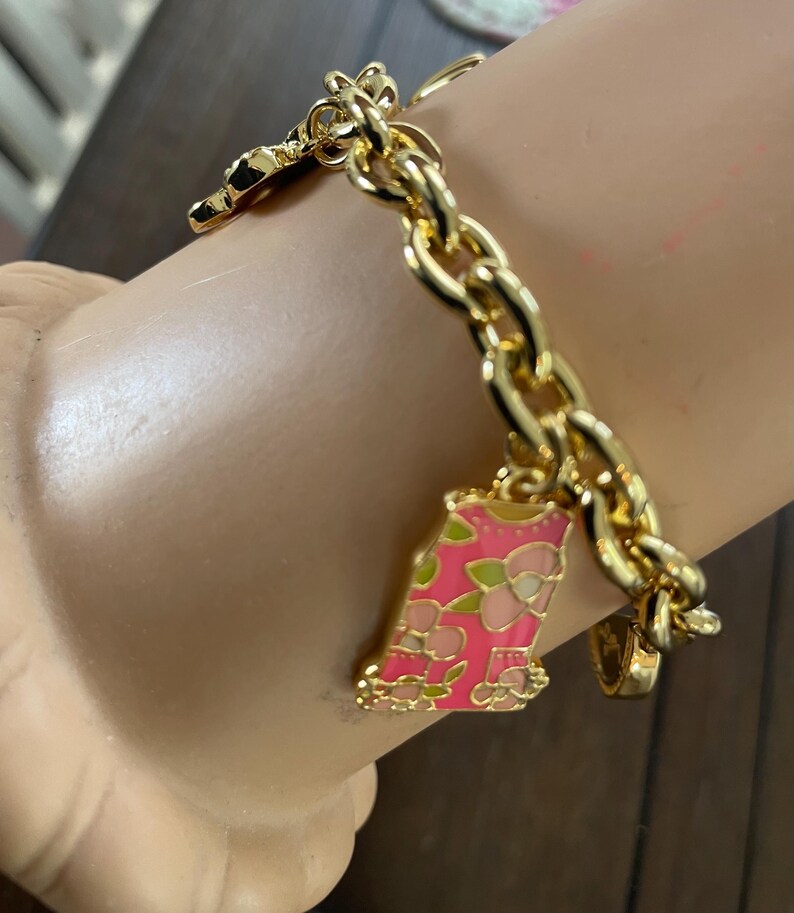 May include: Gold-toned charm bracelet with a chunky chain. The bracelet features several charms, including a pink and green floral-decorated charm. The bracelet is on a light-colored surface.
