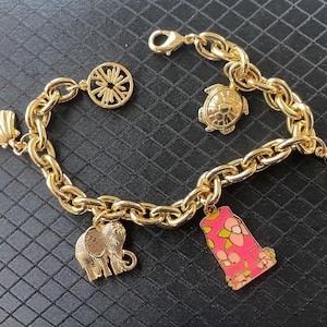 May include: Gold-toned charm bracelet featuring a chunky chain and various charms. Charms include a seashell, a turtle, a slice of fruit, an elephant, a pink and floral design, and a circular design. The bracelet is displayed on a black surface.