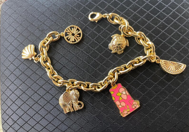 May include: Gold-toned charm bracelet featuring a chunky chain and various charms. Charms include a seashell, a turtle, a slice of fruit, an elephant, a pink and floral design, and a circular design. The bracelet is displayed on a black surface.