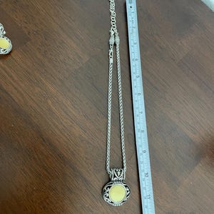 May include: Silver-toned necklace with a round, light yellow pendant. The pendant is approximately 1.25 inches in diameter. The necklace chain is a woven style, and the pendant has an ornate, decorative border.