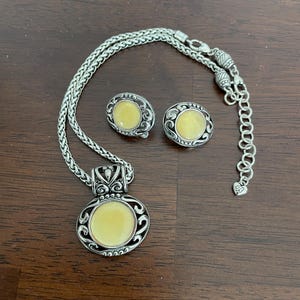 May include: A silver-toned necklace and matching earrings set. The necklace features a pendant with a round, pale yellow center stone, surrounded by an ornate silver design. The earrings have a similar design. The set is displayed on a dark brown surface.