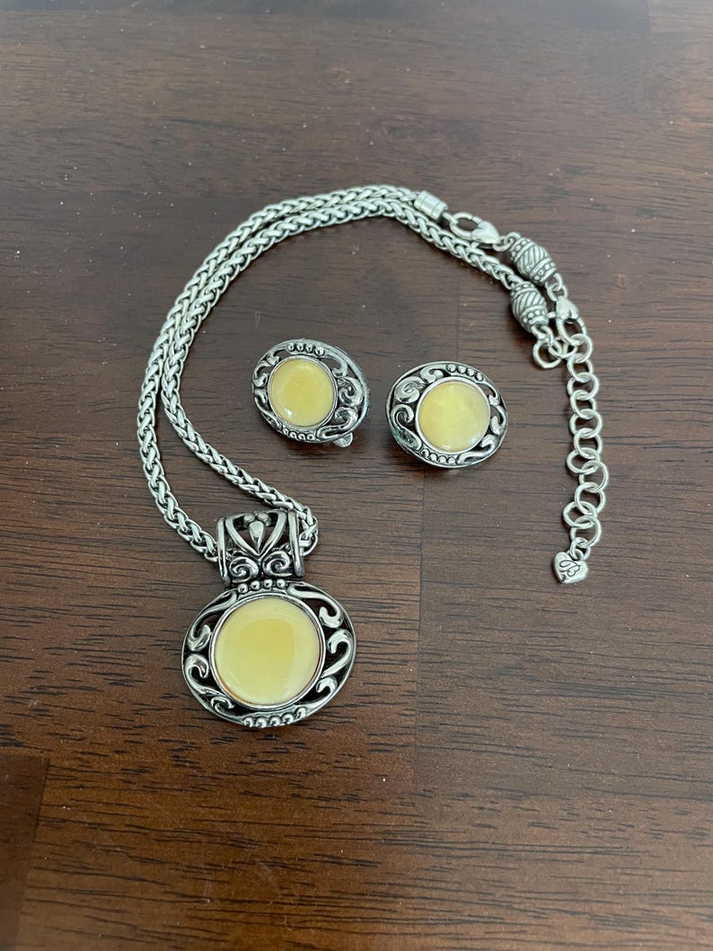 May include: A silver-toned necklace and matching earrings set. The necklace features a pendant with a round, pale yellow center stone, surrounded by an ornate silver design. The earrings have a similar design. The set is displayed on a dark brown surface.
