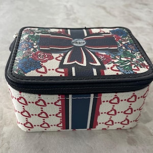 Op de afbeelding: Vierkante sieradendoos met een witte achtergrond en een zwarte ritssluiting. De bovenkant heeft een groot strikdesign met rode, witte en blauwe accenten, omgeven door bloemmotieven. De zijkanten zijn versierd met rode harten.