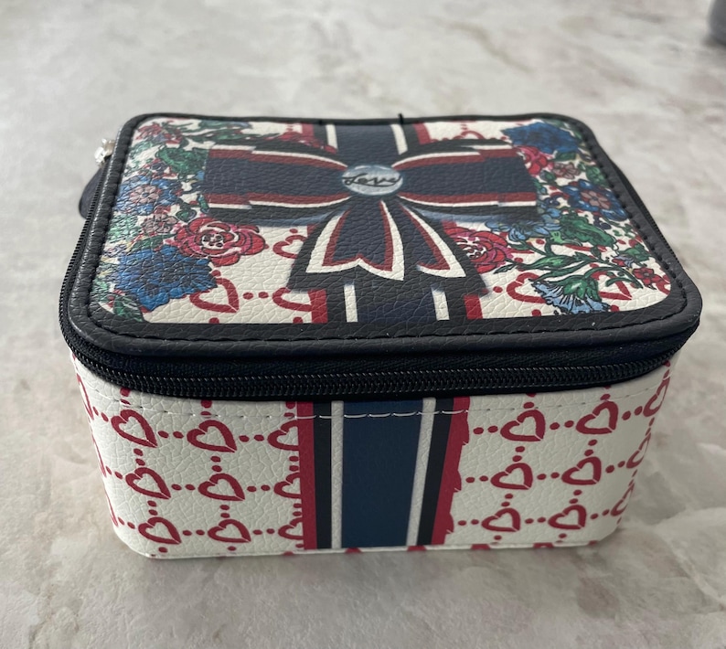 Op de afbeelding: Vierkante sieradendoos met een witte achtergrond en een zwarte ritssluiting. De bovenkant heeft een groot strikdesign met rode, witte en blauwe accenten, omgeven door bloemmotieven. De zijkanten zijn versierd met rode harten.