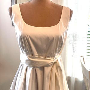 May include: A sleeveless off-white dress featuring a wide scoop neckline and a structured bodice. The waist is accented with a matching fabric sash tied in a knot at the side, leading into a pleated skirt that provides a full, flared silhouette.