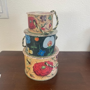 May include: A stack of three cylindrical cardboard boxes with lids, featuring floral and bird illustrations. The top box is cream, the middle is black, and the bottom is yellow, each with a twisted green and cream rope handle attached to the side.