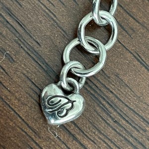 May include: Silver-toned necklace chain with a heart-shaped charm. The charm has a decorative design on it. The chain links are circular and connected. The background is a wooden surface.