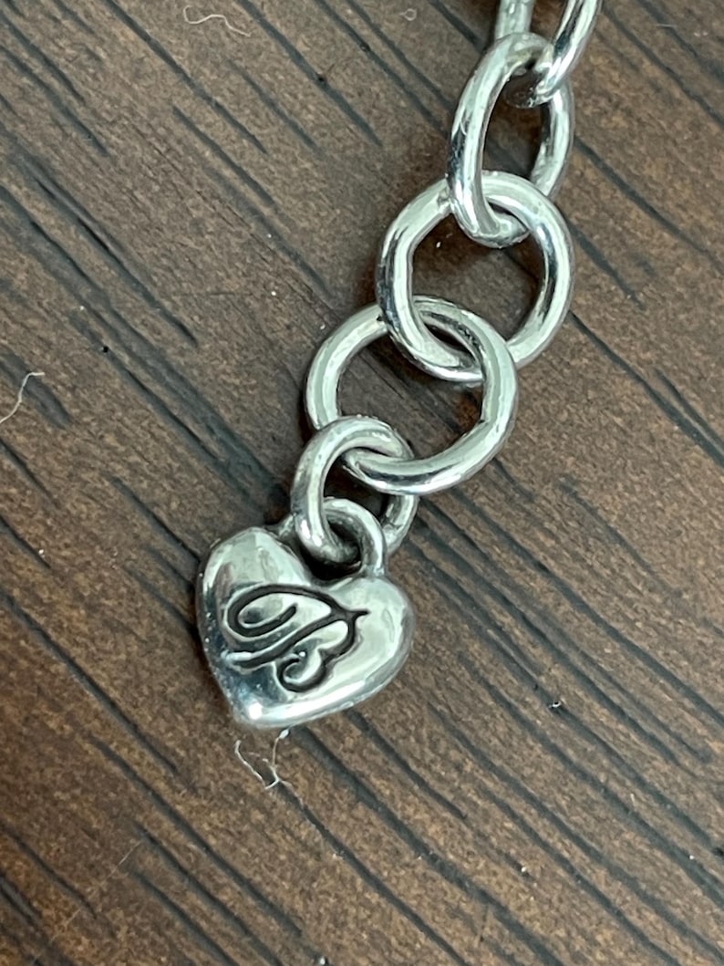 May include: Silver-toned necklace chain with a heart-shaped charm. The charm has a decorative design on it. The chain links are circular and connected. The background is a wooden surface.
