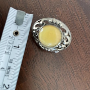 May include: Oval-shaped vintage brooch with a light yellow center stone, surrounded by an ornate silver-tone metal filigree design. The brooch is approximately 1 inch in length.
