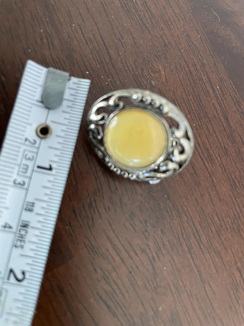 May include: Oval-shaped vintage brooch with a light yellow center stone, surrounded by an ornate silver-tone metal filigree design. The brooch is approximately 1 inch in length.