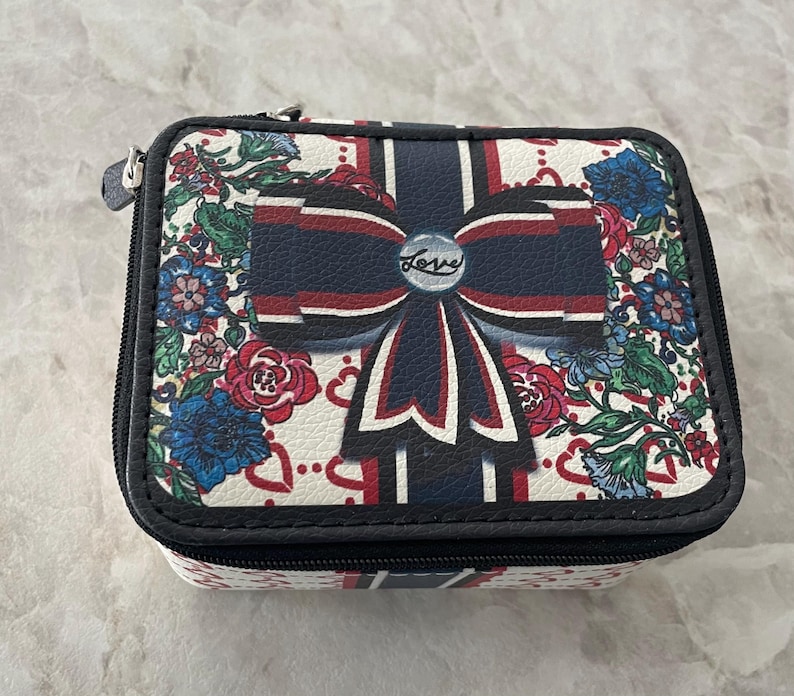 Op de afbeelding: Een rechthoekige sieradendoos met ritssluiting. De koffer heeft een witte achtergrond met een bloemenpatroon en een groot marineblauw strikdesign. Het woord "Love" staat op de strik. De koffer heeft een zwarte rand.