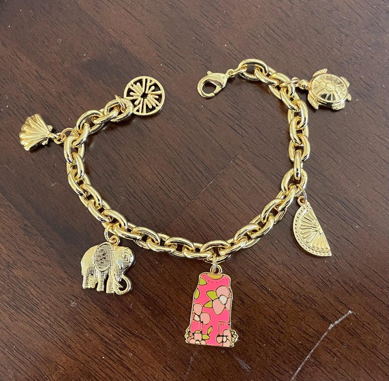 May include: Gold-toned charm bracelet featuring various charms, including a seashell, elephant, turtle, and a pink charm with a floral design. The bracelet has a lobster clasp closure and a chain link design.